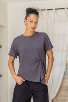 Woman wearing a gray t-shirt and black pants standing in a room with white curtains and wooden door.