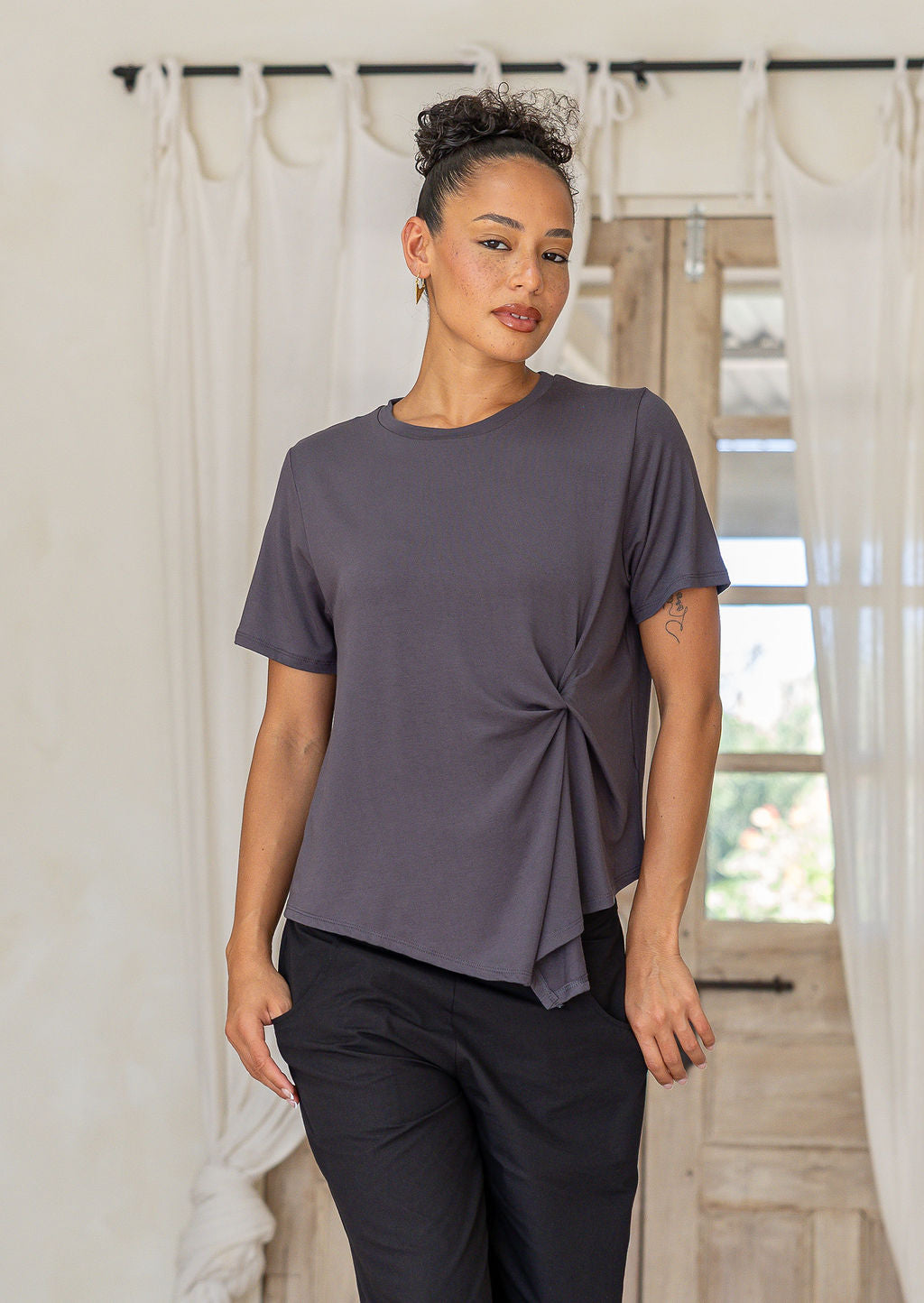 Woman wearing a gray t-shirt with a knot detail and black pants in a room with white curtains.
