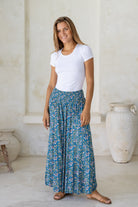 Woman wearing a white t-shirt and blue floral pull on, super wide leg pants, standing in a room with white walls and large vases.