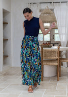 A woman wearing a sleeveless navy blue mock-neck top and long, flowing, wide leg rayon pants in a colourful floral peacock print, and tan sandals