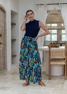 Woman wearing a sleeveless navy blue mock-neck top and high-waisted, wide-leg rayon pants in a multicolour floral and peacock print, in a bright, modern rustic kitchen.