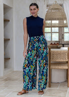 A woman modelling a sleeveless navy top and high-waisted, ruched, wide-leg rayon pants with a vibrant blue, green, and orange floral peacock print