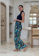 Woman standing side on wearing high-waisted, wide-leg rayon pants with a vibrant blue, green, and yellow floral peacock print, paired with a navy sleeveless top and brown strappy sandals