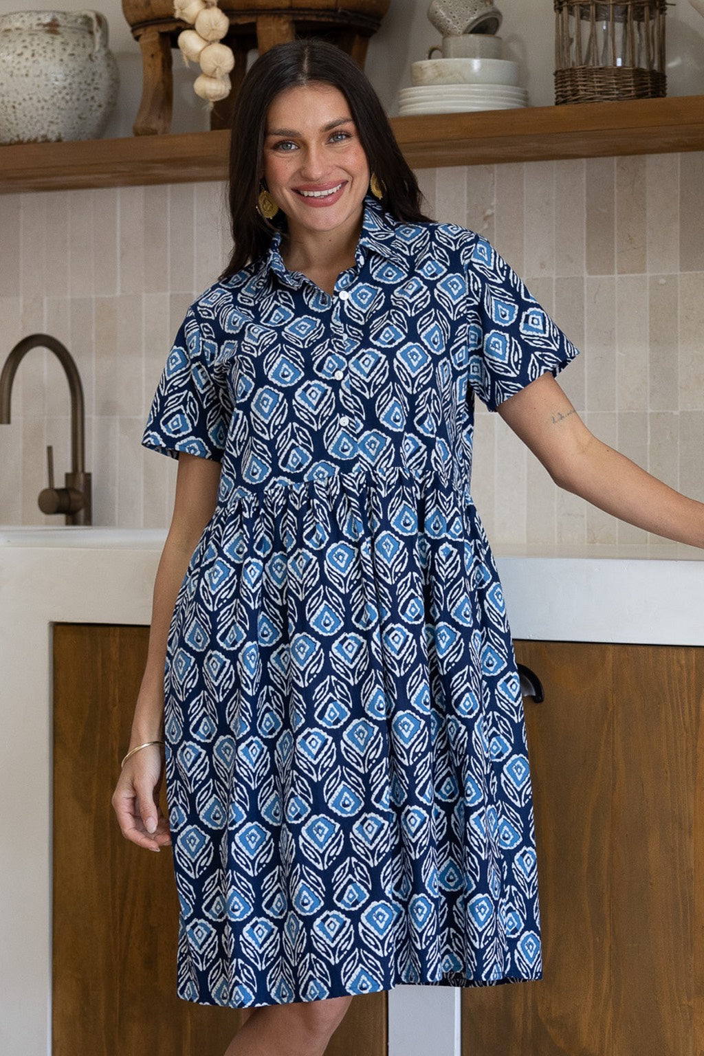 A woman wearing a short-sleeve buttoned bodice relaxed fit cotton dress with a blue, white, and light blue geometric pattern, standing in a kitchen setting.
