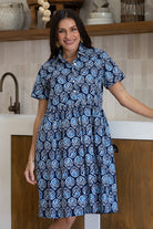 A woman wearing a short-sleeve buttoned bodice relaxed fit cotton dress with a blue, white, and light blue geometric pattern, standing in a kitchen setting.
