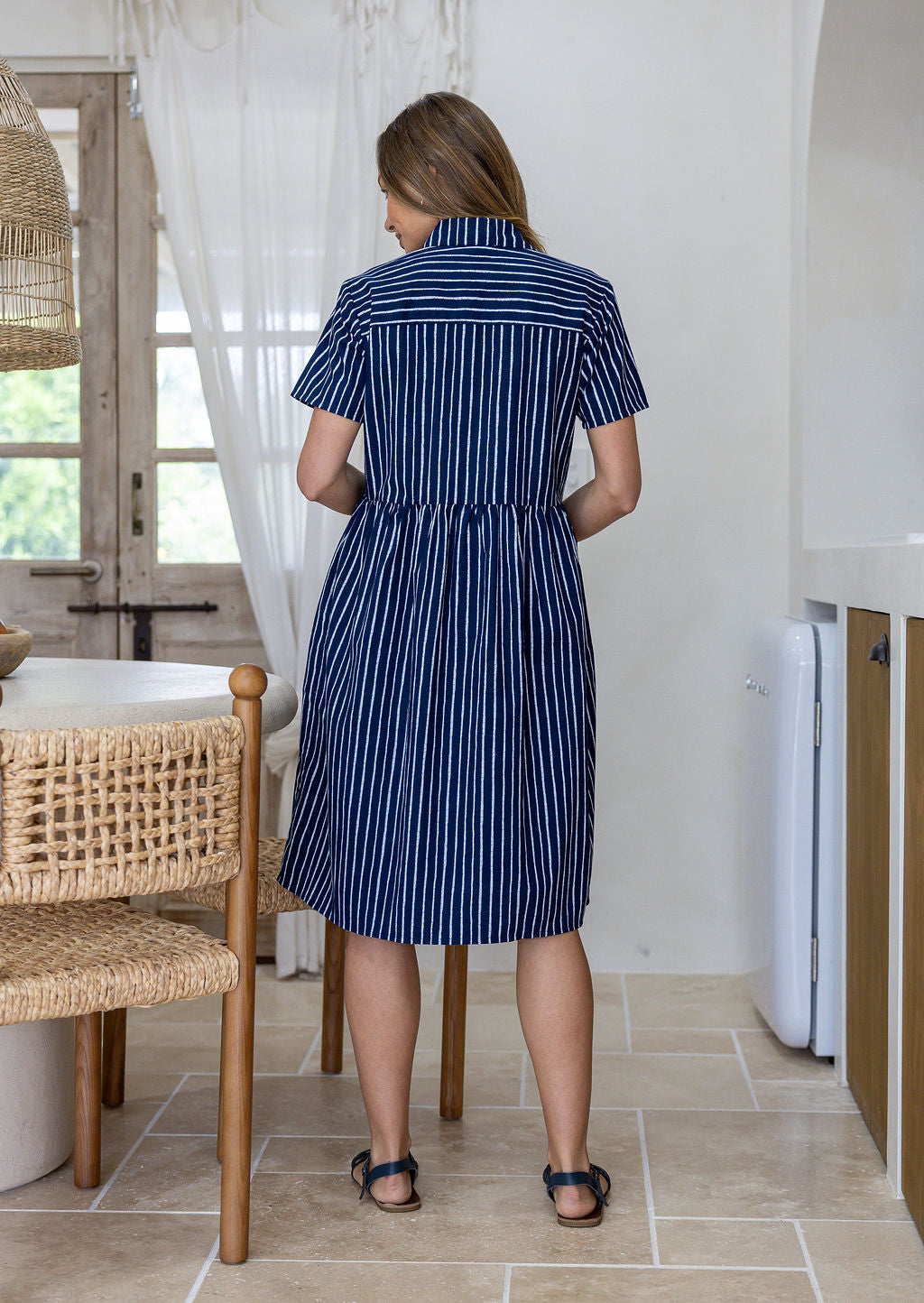 Woman standing backwards wearing a 100% cotton relaxed fit collared short sleeve dress with hidden side pockets.