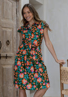 Woman in a olive green based floral cotton dress with cap sleeves, buttoned bodice, pockets, box pleats at the waist and a fabric waist tie.
