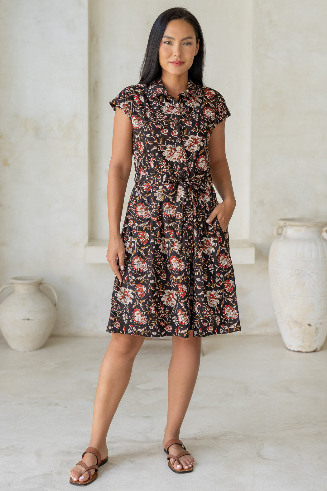 Model stands with one hand in pocket wearing a 100% cotton knee length shirt style retro floral dress in black based indian traditional print.