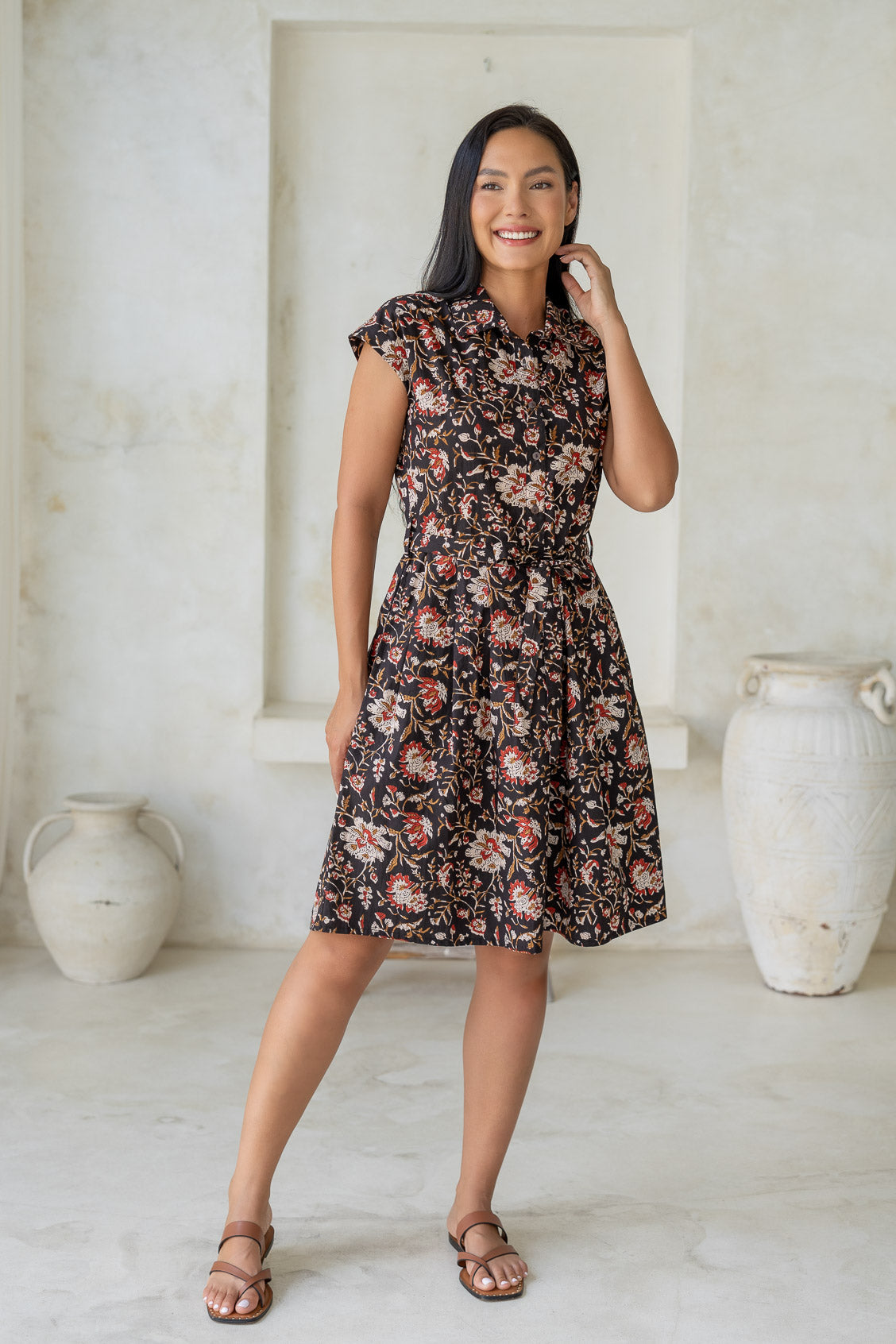 Woman wearing a knee length shirt style retro floral dress in black based floral print, standing in a room with white walls and large vases.