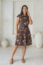 Woman wearing a knee length shirt style retro floral dress in black based floral print, standing in a room with white walls and large vases.