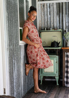 Woman in a gentle cream based floral cotton dress with cap sleeves, buttoned bodice, pockets, box pleats at the waist and a fabric waist tie.