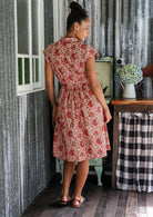 Woman stands backwards wearing a gentle cream based floral cotton dress with cap sleeves, buttoned bodice, pockets, box pleats at the waist and a fabric waist tie.