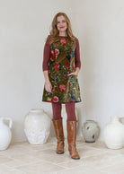 Woman wearing a moss green based multi-coloured floral cotton velvet tunic with boat neck and one hand in pocket.