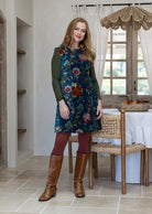 Woman wearing a cotton velvet sleeveless tunic in a teal based multi-coloured floral print with brown boots. 