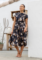 Woman poses in 1950's style short sleeved, button through cotton dress with high round neckline in black based floral print.