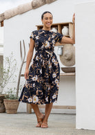 Woman wears 100% cotton button through midi length dress with short sleeves and pockets in floral print on a black base.