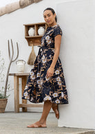 Woman wearing a black floral 100% cotton dress standing against a white wall with a wooden shelf in the background.