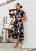Woman wears black based floral printed cotton button through cotton dress with hidden side pockets, short sleeves and a detachable waist tie.