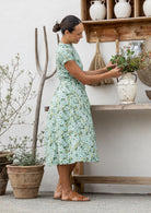 Woman stands sideways wearing a 1950's style short sleeved, button through cotton dress with high round neckline in sage green based floral print.