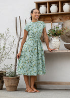 Woman wears retro style cotton button through midi length dress with high round neckline, short sleeves and a removable waist tie.