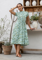 Woman wears 100% cotton button through midi length dress with short sleeves and pockets in floral print on a sage green base.