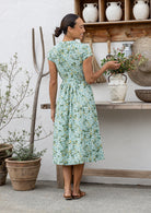 Woman stands backwards wearing a sage green based floral printed cotton button through cotton dress with hidden side pockets, short sleeves and a detachable waist tie.