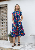 Woman wears 100% cotton button through midi length dress with short sleeves and pockets in floral print on a royal blue base.