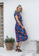Woman stands sideways wearing a royal blue based floral printed cotton button through cotton dress with hidden side pockets, short sleeves and a detachable waist tie.