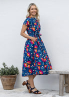 Woman stands sideways wearing a cotton button through cotton dress with hidden side pockets, short sleeves and a detachable waist tie.