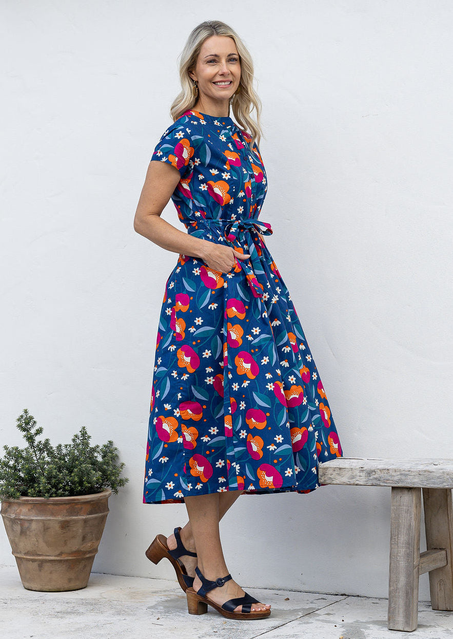 Woman stands sideways wearing a cotton button through cotton dress with hidden side pockets, short sleeves and a detachable waist tie.