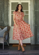 Woman in a floral dress standing in front of a white door with a wicker chair and cushions.