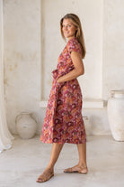 Women stands in side pose with hands in pockets wearing floral printed midi length dress with high round neckline, short sleeves and pockets.