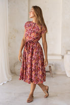 Woman stands slightly sideways wearing a cotton short sleeved dress with detachable waist tie and button through front in a warm pinkish toned floral print.