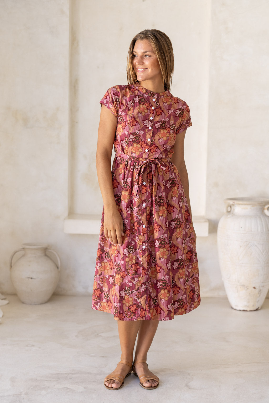 Woman poses in retro style cotton button through dress with short sleeves, high round neckline and a fabric waist tie in a warm toned floral print.