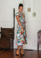 Woman wearing a colorful floral dress standing in a room with wooden furniture.