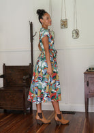 Woman stands sideways wearing a colourful floral printed cotton button through cotton dress with hidden side pockets, short sleeves and a detachable waist tie.