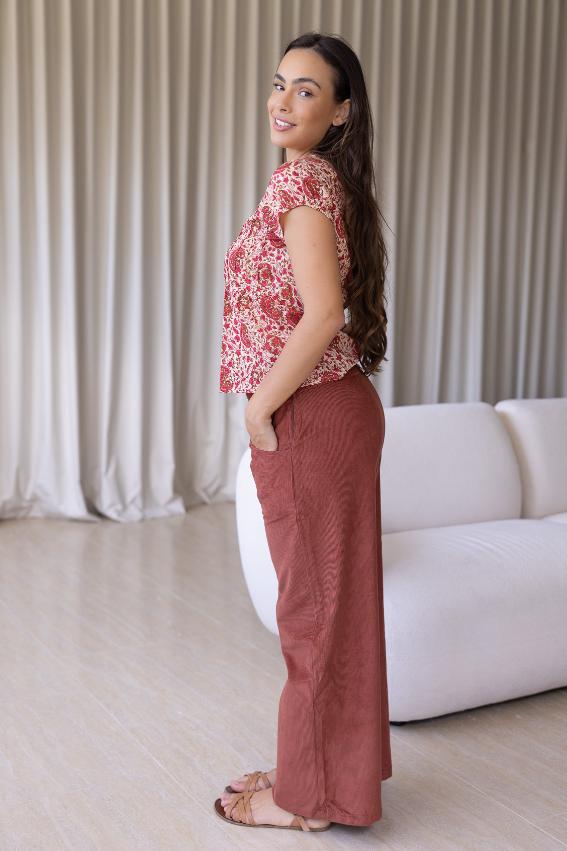 Woman stands backwards wearing a gentle cream based floral print cotton top paired with plain pants.