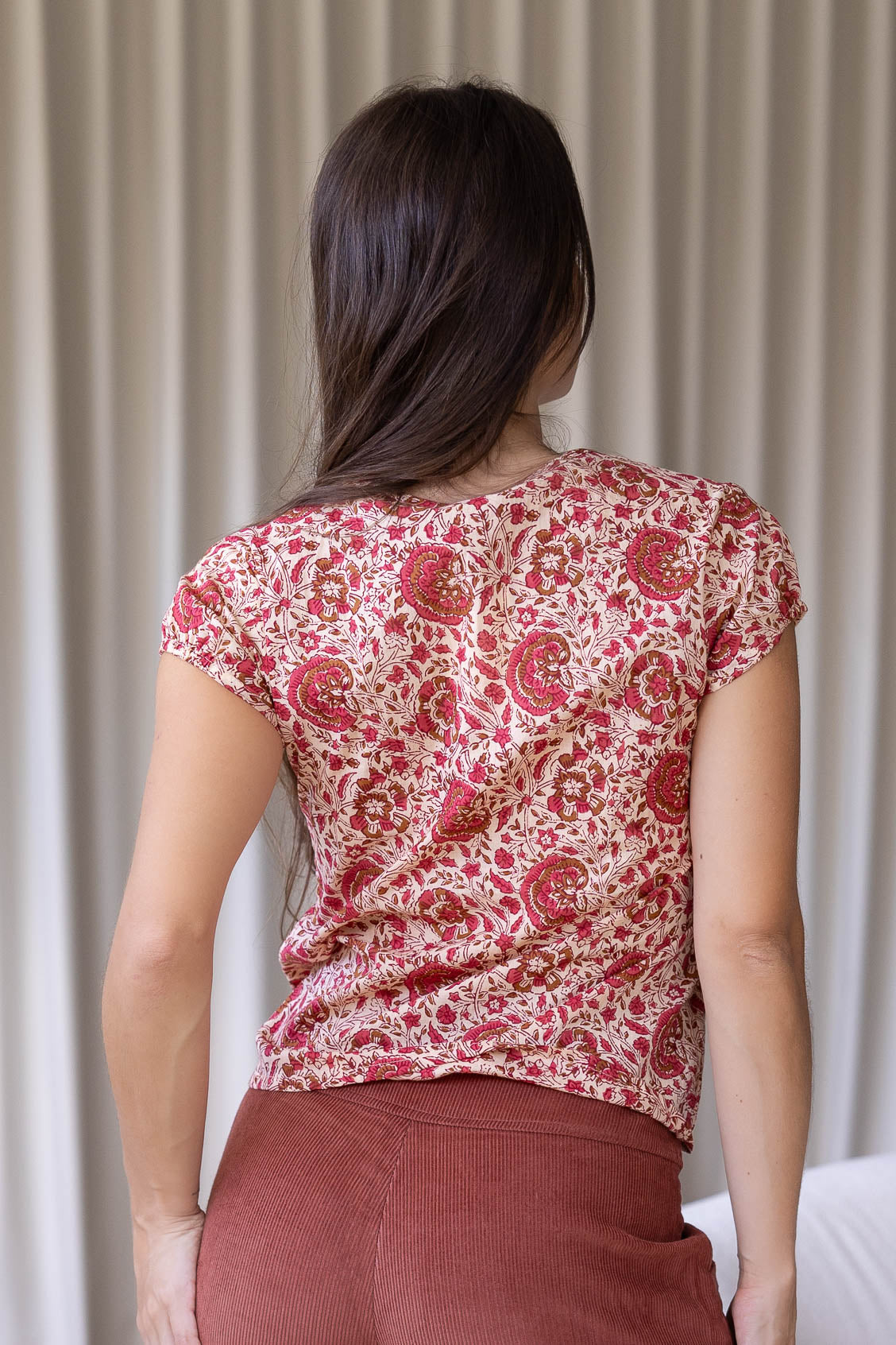 Woman stands backwards wearing a gentle cream based floral print cotton top standing against a white wall.