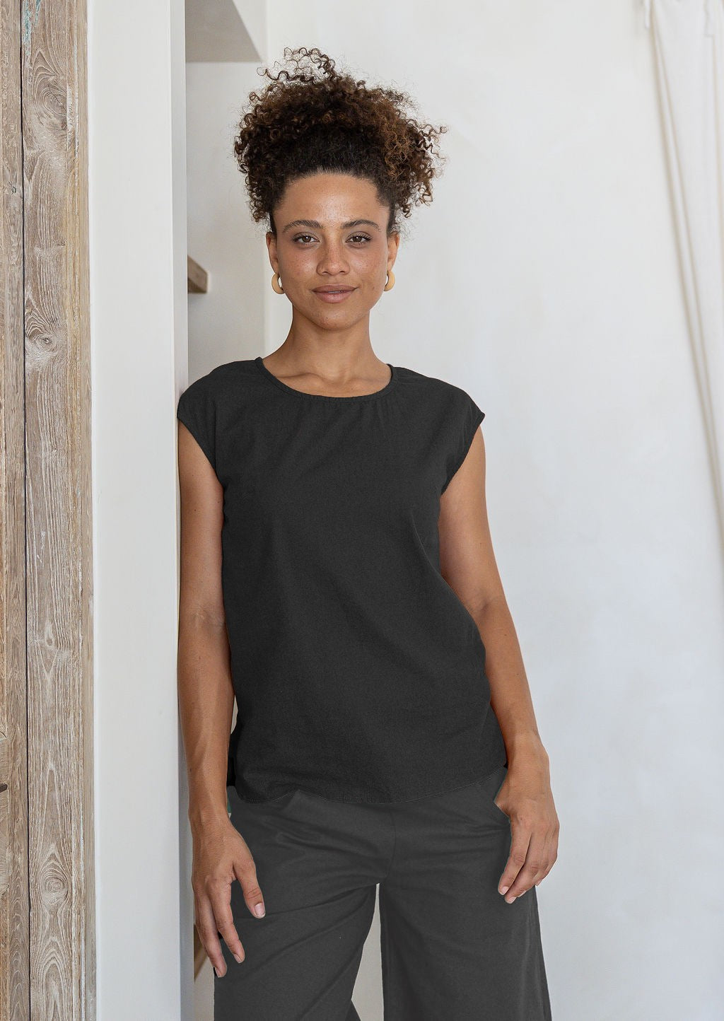 Woman wearing a black cotton sleeveless top and pants against a white wall.