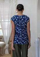 Woman stands backwards in a navy based floral sleeveless cotton top with button closure at the back paired with plain pants  