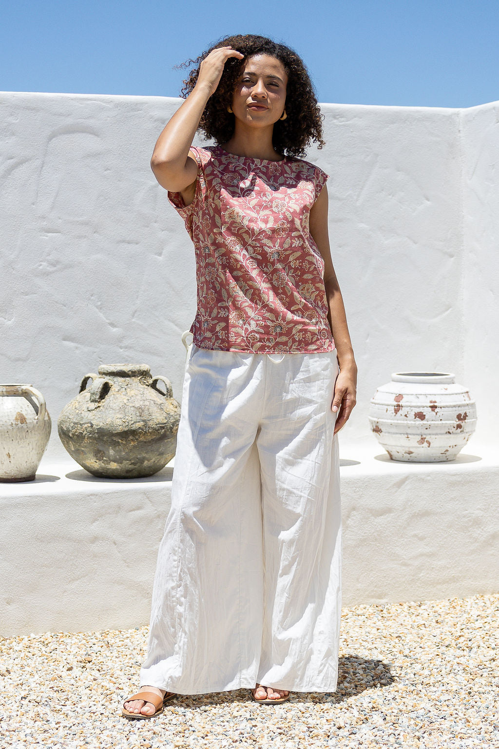 Woman in a floral warm pink based cotton top and white pants standing in front of a white wall with decorative pots.