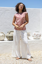 Woman wearing a warm pink based floral cotton top and white pants standing in front of a white wall