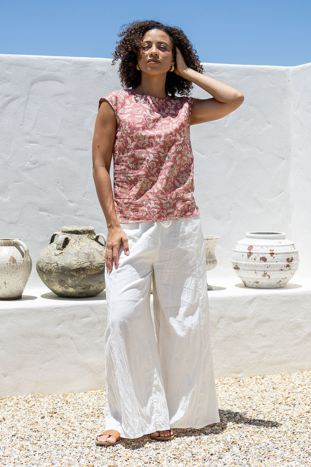 Woman wearing a pink sleeveless cotton top and white pants standing in front of a white wall with pots.