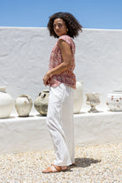 Woman in a warm pink based floral sleeveless cotton top and white pants standing in front of a white wall