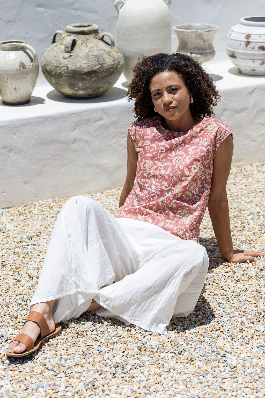 Woman sitting on pebbles wearing a warm pink floral cotton sleeveless top and white pants