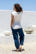 Back view of woman in a white sleeveless cotton top with a button closure at the nape of neck and a box pleat in the centre of the back, paired with blue pants