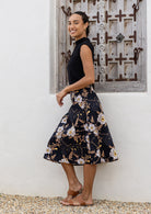 Woman standing sideways wearing a black top and black floral cotton skirt against a wooden window,