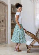 Woman in a white top and floral skirt standing in a room with wooden door and chair.