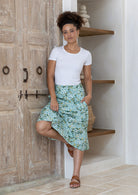 Woman wearing a white top and sage green floral skirt standing in a room with wooden door and shelves.