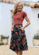 Woman wearing a basic rust top and black floral a-line cotton skirt with side zipper and hands in pockets.